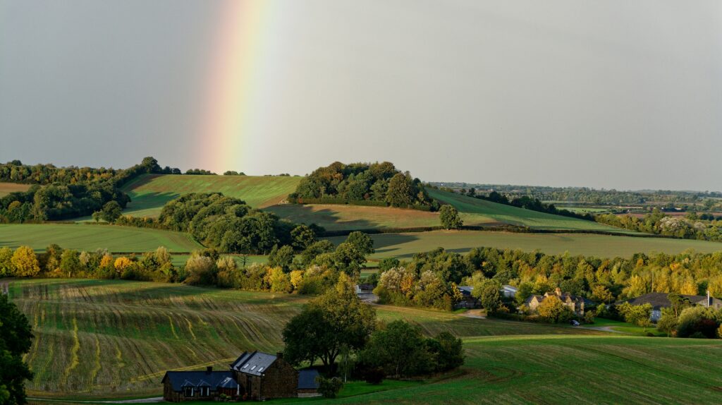 Rural setting with houses, farm buildings, fields, hills and a rainbow.